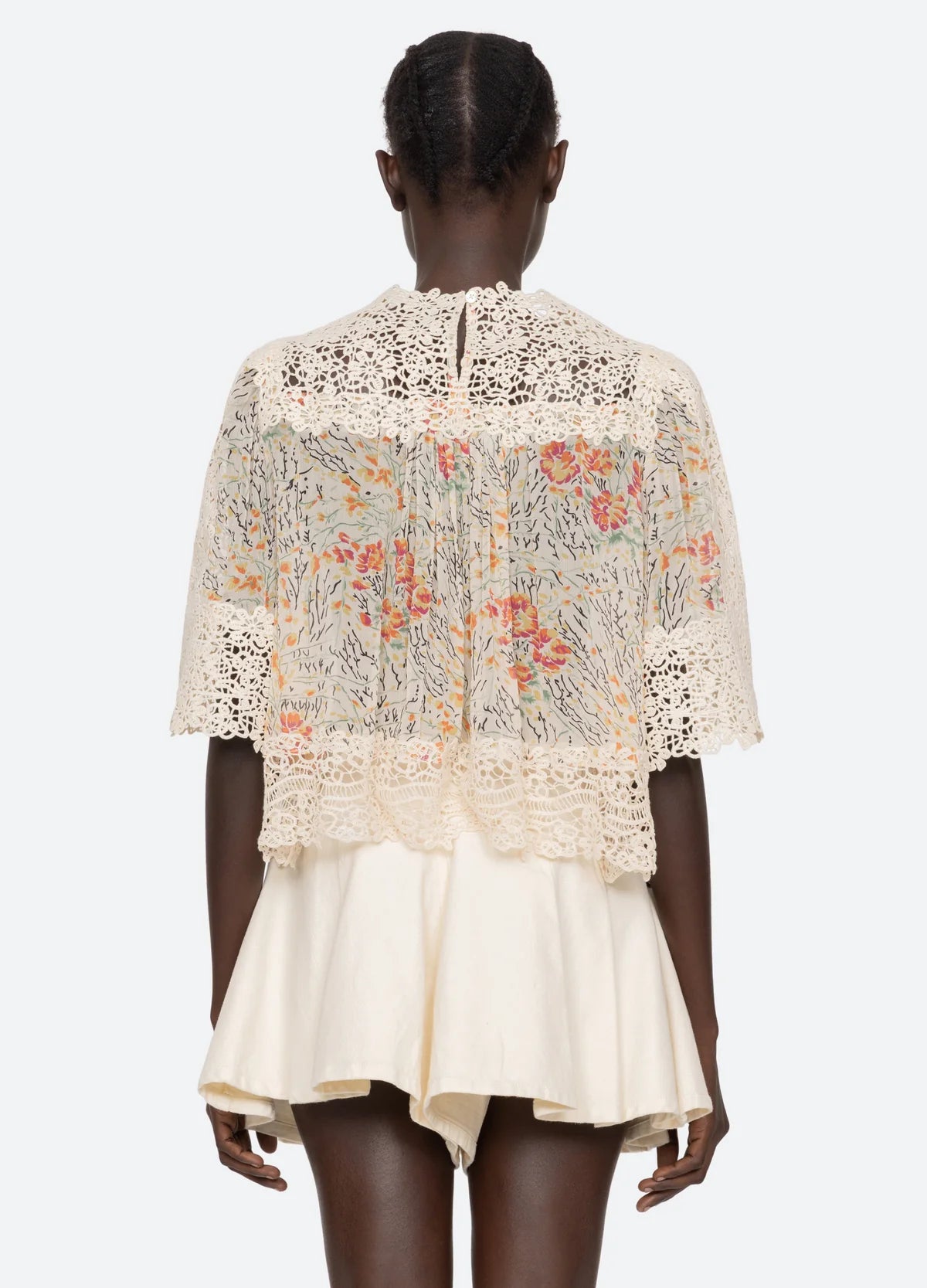A person facing away wears the Luisa Print SS Top by Sea New York, a relaxed fit sheer lace top with colorful floral embroidery and cream crochet details over a short cream skirt. The top has scalloped edges. The background is plain white.