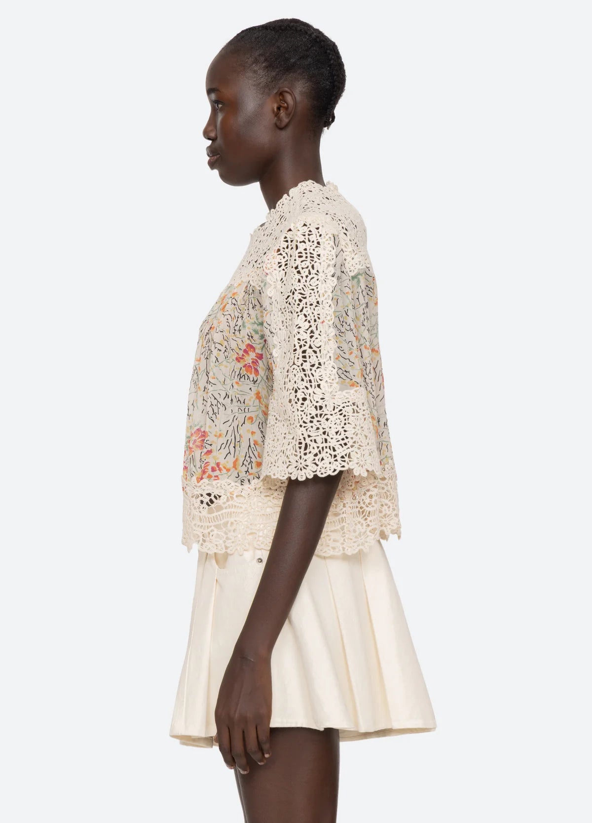 A woman stands in profile wearing the Sea New York Luisa Print SS Top, a relaxed cream crochet lace piece with floral patterns and flared short sleeves, paired with a cream pleated skirt against a light background.