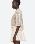 A woman stands in profile wearing the Sea New York Luisa Print SS Top, a relaxed cream crochet lace piece with floral patterns and flared short sleeves, paired with a cream pleated skirt against a light background.