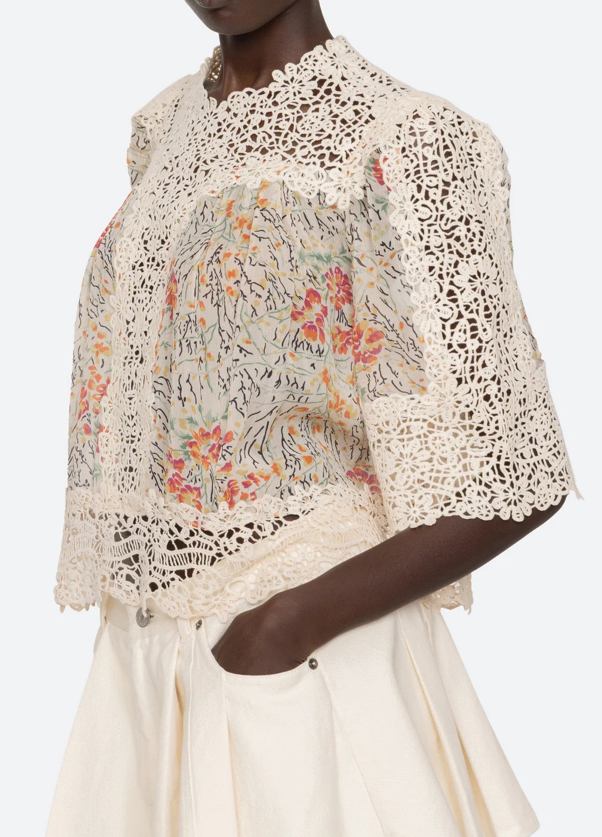A person wears the Luisa Print SS Top by Sea New York, a cream-colored relaxed-fit lace crochet piece with floral embroidery and short sleeves, paired with cream high-waisted shorts. Their hand is in their pocket.