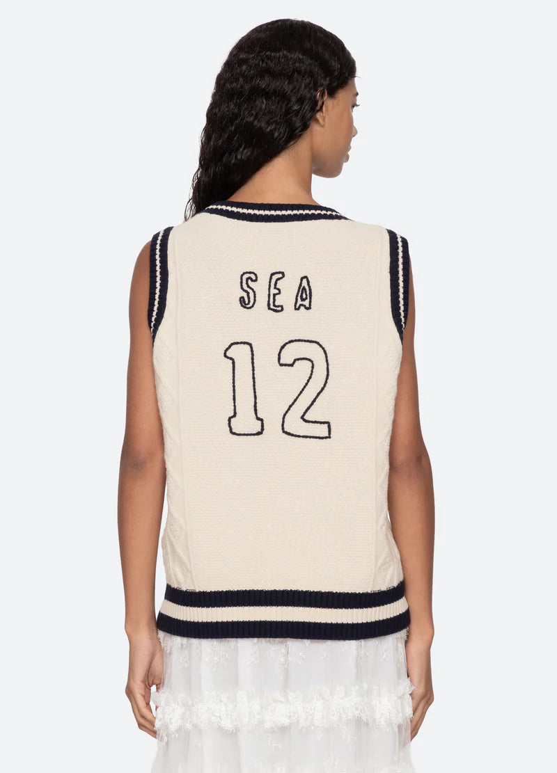 A woman with wavy dark hair faces away, wearing the Sea New York Maren Knit Tank—a sleeveless cream cable-knit vest with navy trim and "SEA 12" in navy on the back—paired with a white skirt.