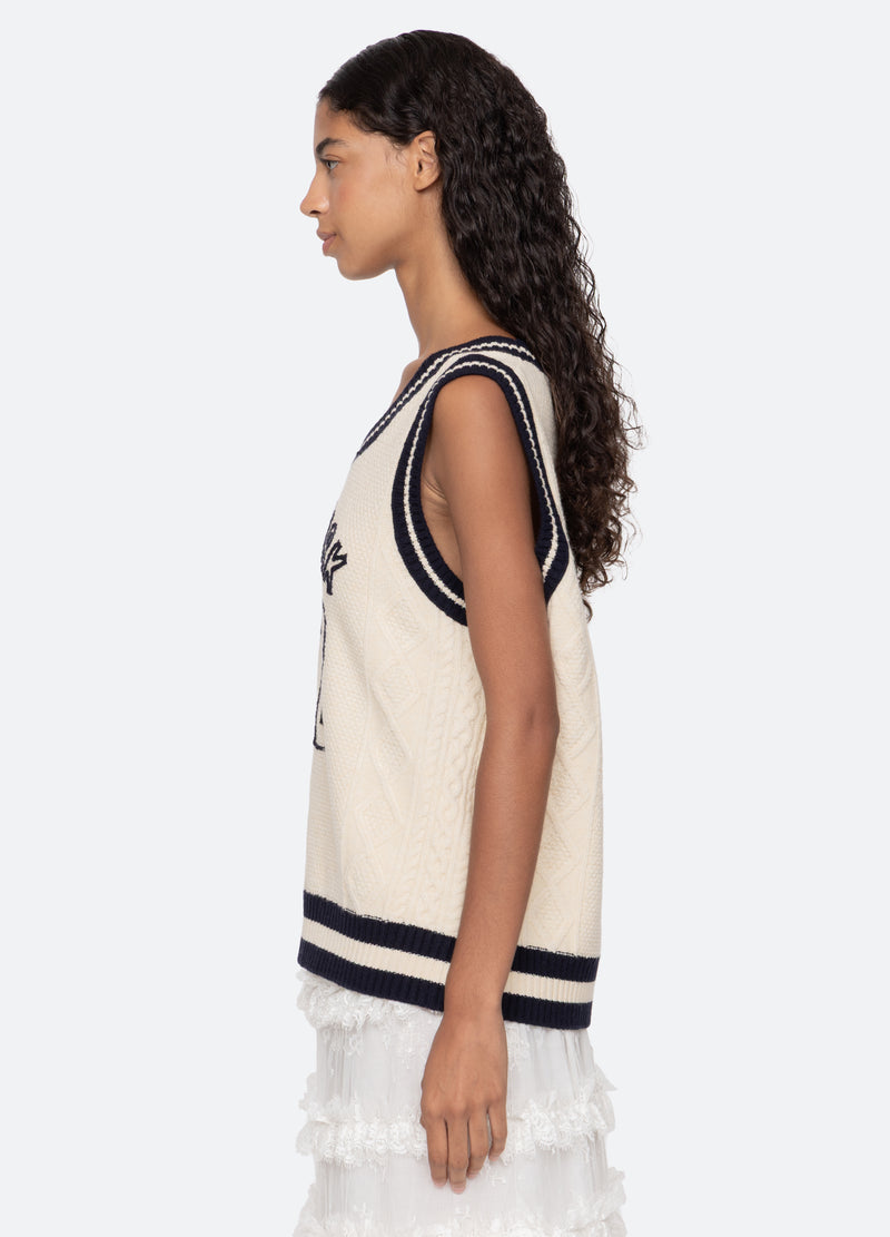 A woman with long, curly hair is shown in profile wearing the Sea New York Maren Knit Tank—a cream-colored, cable-knit vest with black trim—paired with a white lace skirt against a plain light background.