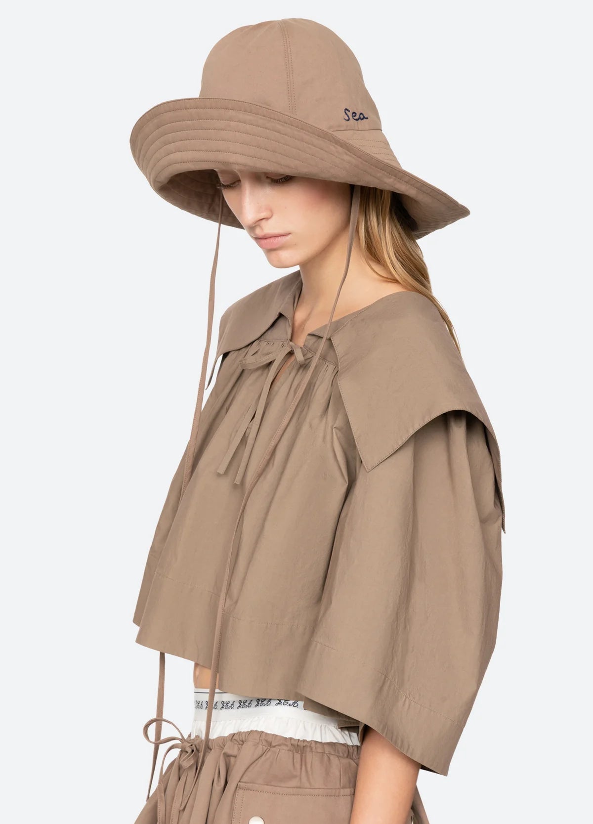 A woman in a loose taupe top and matching drawstring shorts looks down. She wears the Sea New York Lois Twill Hat, an oversized sun hat with long chin ties and "Sea" embroidery, for effortless style.