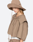 A woman in a loose taupe top and matching drawstring shorts looks down. She wears the Sea New York Lois Twill Hat, an oversized sun hat with long chin ties and "Sea" embroidery, for effortless style.