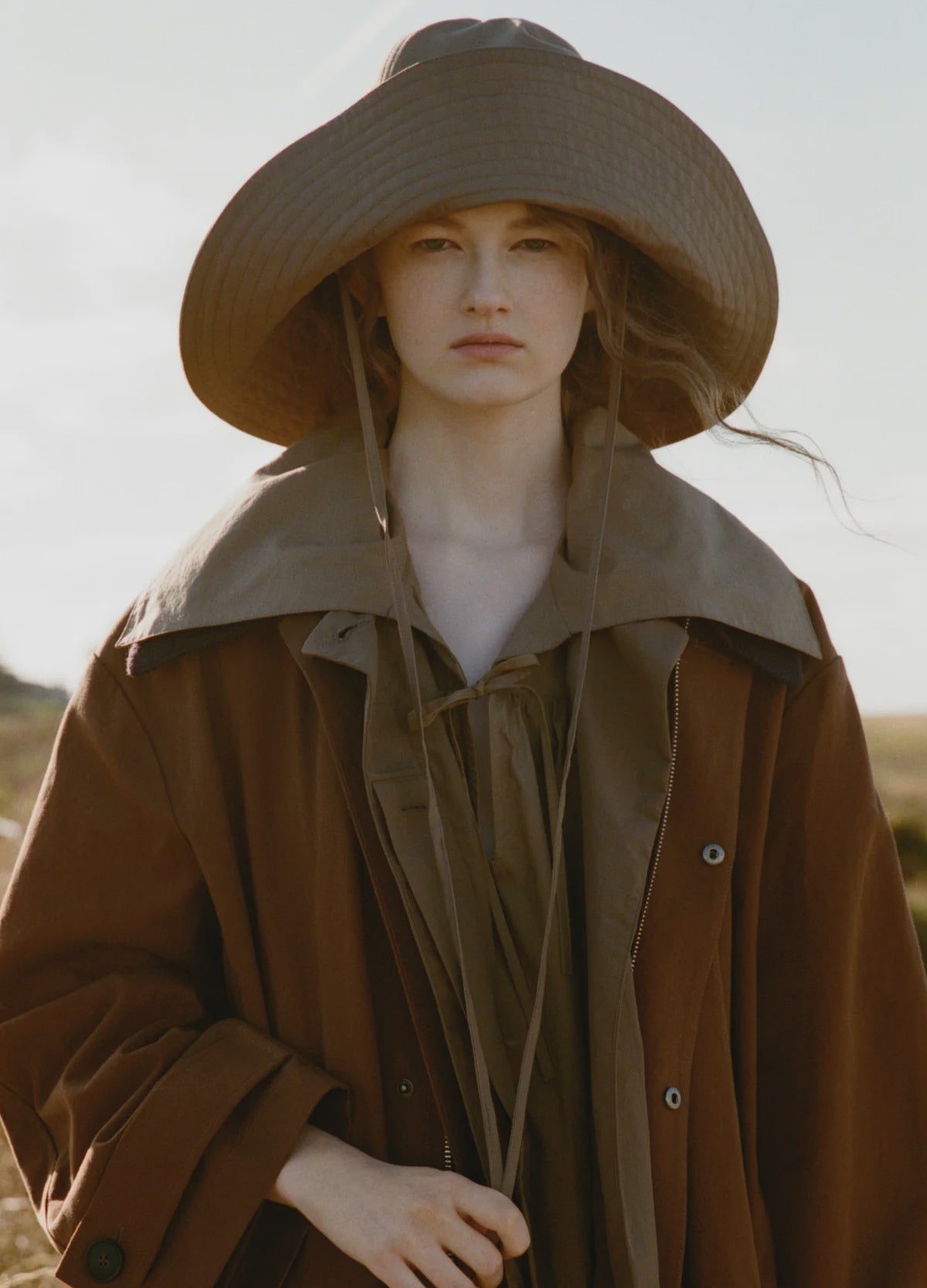 A person wears the Lois Twill Hat by Sea New York and a loose brown coat, standing outdoors in a natural, softly blurred setting and looking directly at the camera with a neutral expression.