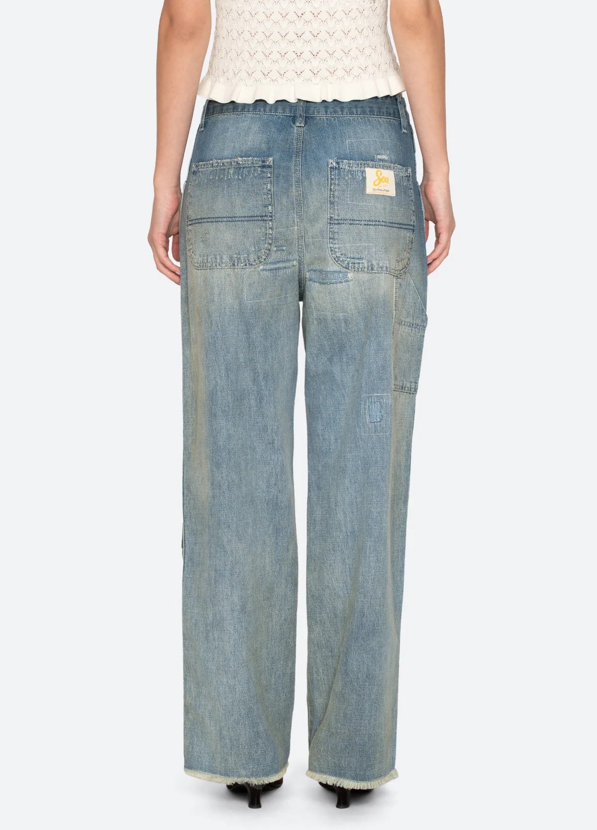 A person wearing Sea New York's Claira Mended Jeans in relaxed fit light blue denim, seen from the back with visible pockets and a patch on one pocket, paired with a cream knitted top.