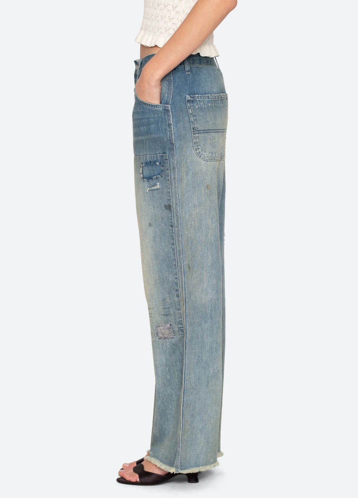 A person in Sea New York’s Claira Mended Jeans—relaxed fit with distressed patchwork and frayed hems—stands sideways, one hand in pocket, paired with open-toe heels.