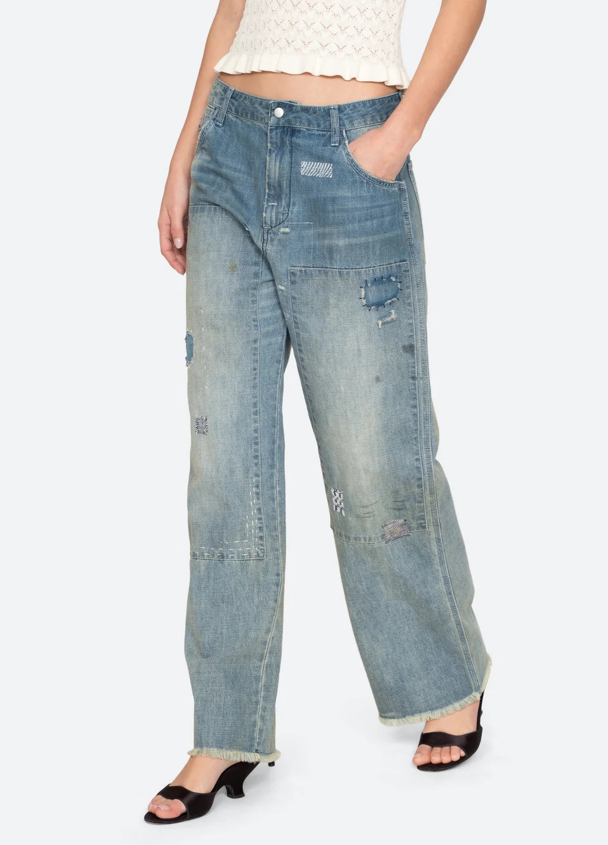 A person wearing Sea New York's Claira Mended Jeans—relaxed fit, wide-leg, light-wash with patchwork and raw hem—styled with a textured white crop top and black open-toe heels.