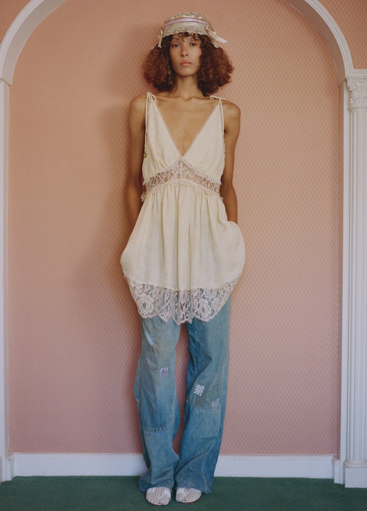 A person with curly hair wears Claira Mended Jeans by Sea New York, a lacy cream dress, sandals, and a decorative headpiece, posing against a pink wall framed by a white arch.