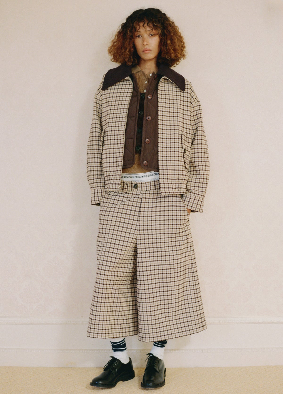 Wearing the Savannah Plaid Culotte by Sea New York, a person with curly hair pairs it with a loose checked jacket, brown shirt, visible boxer-style waistband, striped socks, and black shoes while standing against a pale wall.