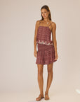 A woman poses against a light background, wearing natalie martin’s Chelsea Mini Skirt with an elastic waistband and a matching sleeveless maroon-and-white patterned rayon top with lace trim. She accessorizes with sandals and minimal jewelry.