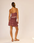 A woman with her hair tied back stands on a plain light background, wearing the natalie martin Chelsea Mini Skirt—featuring a sleeveless, patterned red design with an elastic waistband and ruffled mini hem.