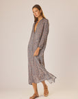 A woman stands in a studio wearing the natalie martin MB Dress—a patterned midi with long sleeves, straight fit, and deep V-neck—paired with flat sandals. She gazes slightly to the side with a neutral expression.