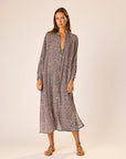 A woman stands against a plain background wearing the natalie martin MB Dress, a loose, straight-fit v-neck midi with a colorful geometric pattern, paired with brown sandals.