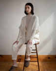 A woman with long dark hair sits calmly on a wooden stool in a softly lit room, wearing LaTierra’s Nube Vest over matching off-white pants and brown slippers with white trim. The wall and floor are plain, complementing her neutral expression.