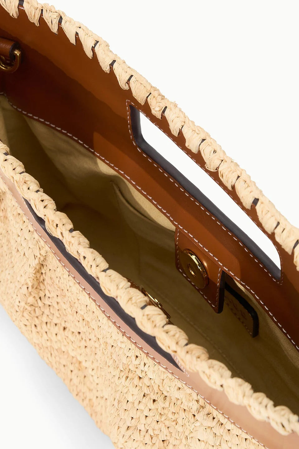 A close-up of the Staud Lena Raffia Clutch shows its woven straw design, tan leather trim and handles, open top with built-in handles, and snap button closure.