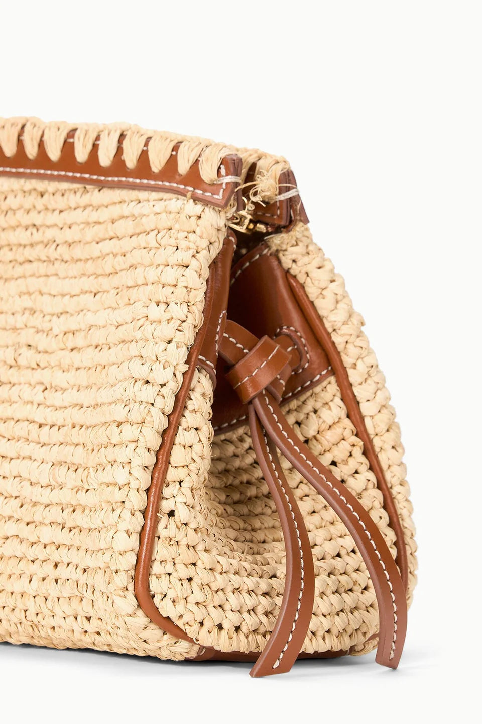 A close-up of the Staud Maude Convertible Clutch in Natural, showcasing woven raffia, brown leather trim, decorative stitching, a leather shoulder strap, and tie accents on the side.