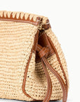 A close-up of the Staud Maude Convertible Clutch in Natural, showcasing woven raffia, brown leather trim, decorative stitching, a leather shoulder strap, and tie accents on the side.