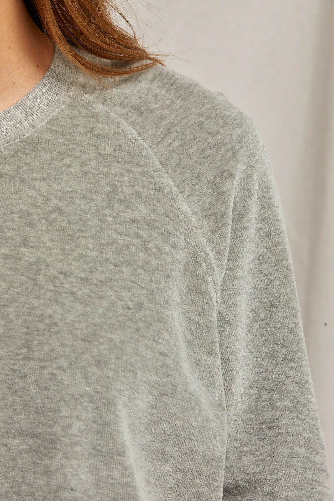 Close-up of a person wearing the gray Stella Velour Shrunken Raglan Sweatshirt from Perfectwhitetee, showcasing the textured fabric and raglan seaming. The image emphasizes the upper section, highlighting the shoulder and part of the sleeve, while a neutral light background enhances the luxurious appearance of this soft garment.