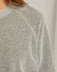 Close-up of a person wearing the gray Stella Velour Shrunken Raglan Sweatshirt from Perfectwhitetee, showcasing the textured fabric and raglan seaming. The image emphasizes the upper section, highlighting the shoulder and part of the sleeve, while a neutral light background enhances the luxurious appearance of this soft garment.