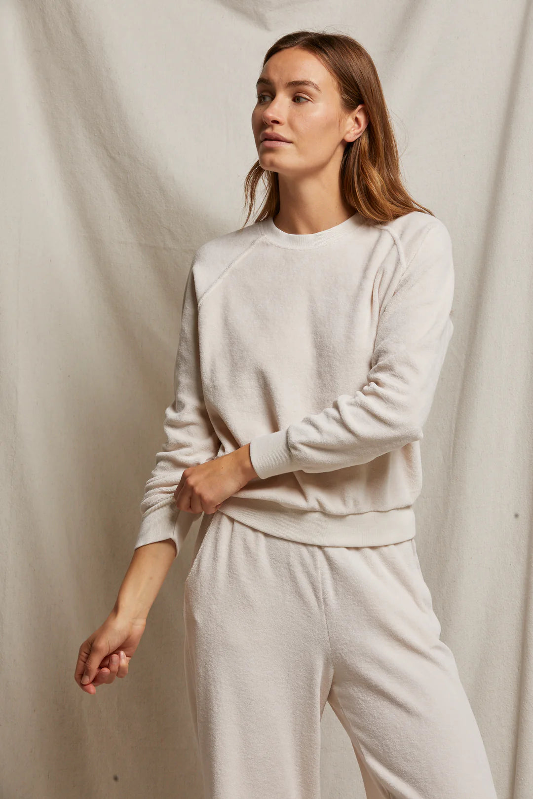 A person models the Stella Velour Shrunken Raglan Sweatshirt by Perfectwhitetee, styled with matching cream-colored sweatpants in a relaxed fit. They stand against a light fabric backdrop, showcasing long hair and a neutral expression as one hand rests on their opposite wrist.