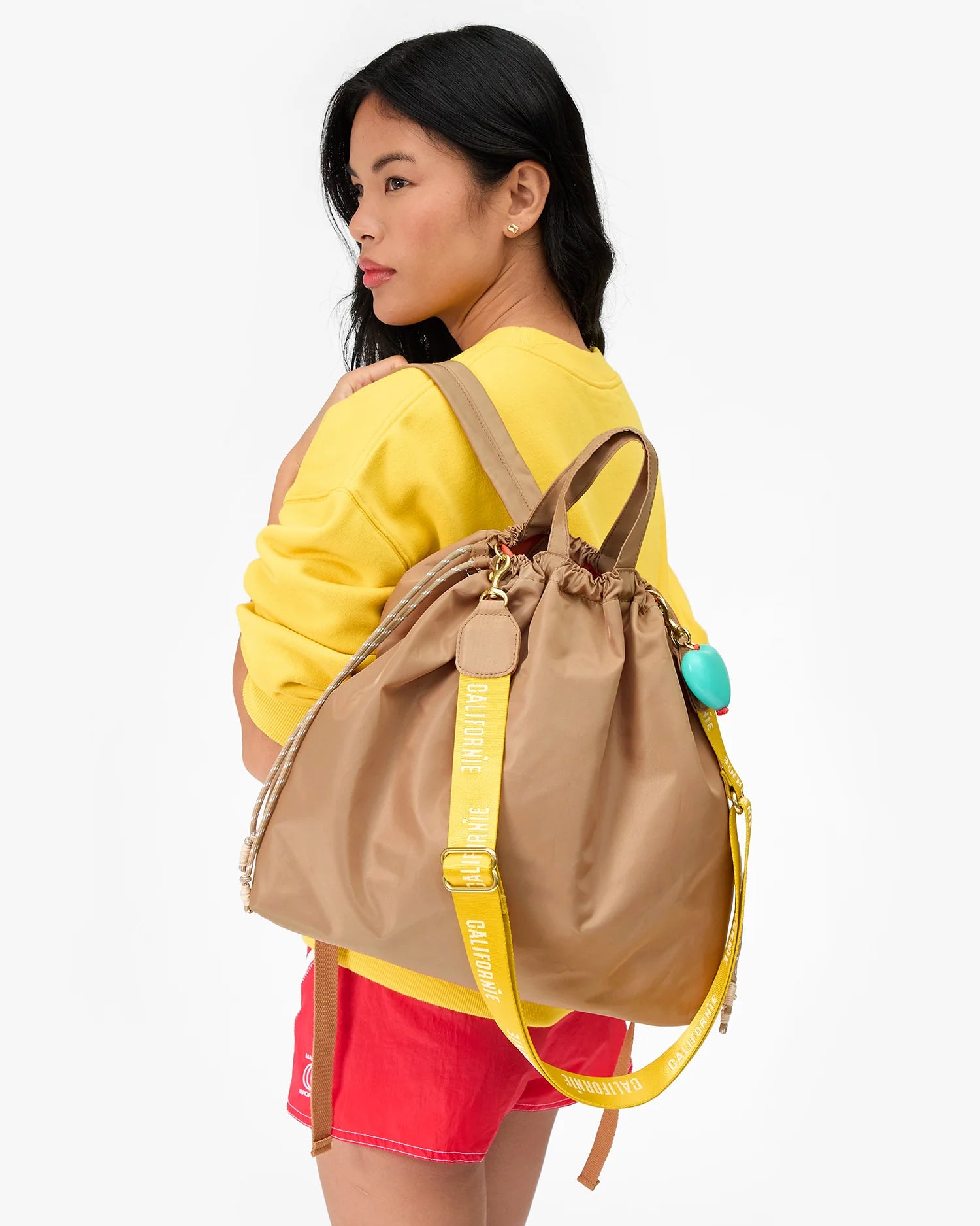 A woman with long dark hair, wearing a yellow sweatshirt and red shorts, carries the Clare Vivier Sac Tout Backpack—a large tan recycled nylon bag with yellow straps and a turquoise accessory—over her shoulder.