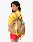 A woman with long dark hair, wearing a yellow sweatshirt and red shorts, carries the Clare Vivier Sac Tout Backpack—a large tan recycled nylon bag with yellow straps and a turquoise accessory—over her shoulder.