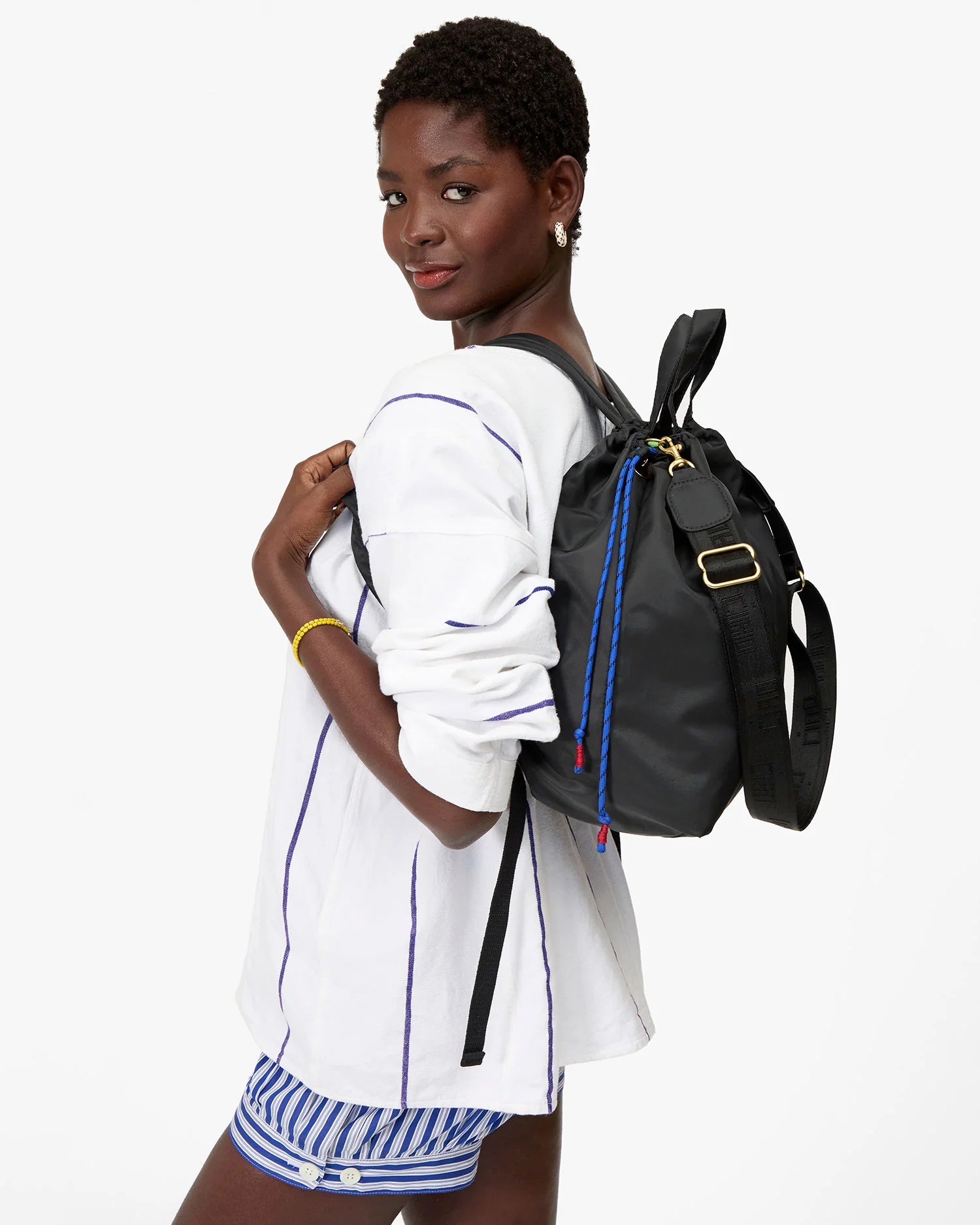 A woman wears a white long-sleeve shirt with blue stripes and matching shorts, posing confidently with the Clare Vivier Sac Tout Backpack over her shoulder against a plain white background.
