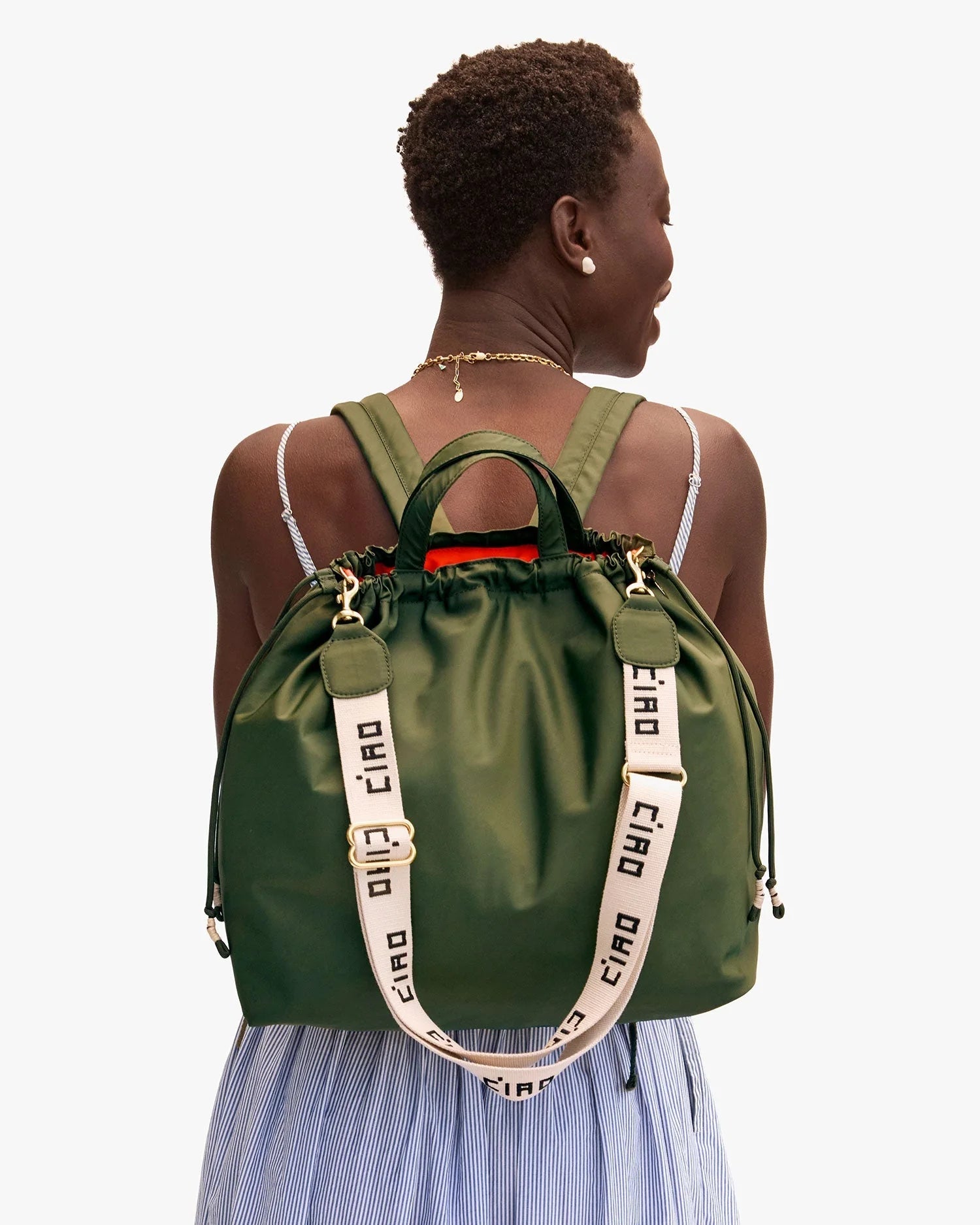 A person with short hair, in a sleeveless light blue and white striped dress, carries Clare Vivier’s Sac Tout Backpack in olive recycled nylon with cream “ciao” straps, gold hardware, and black cords.