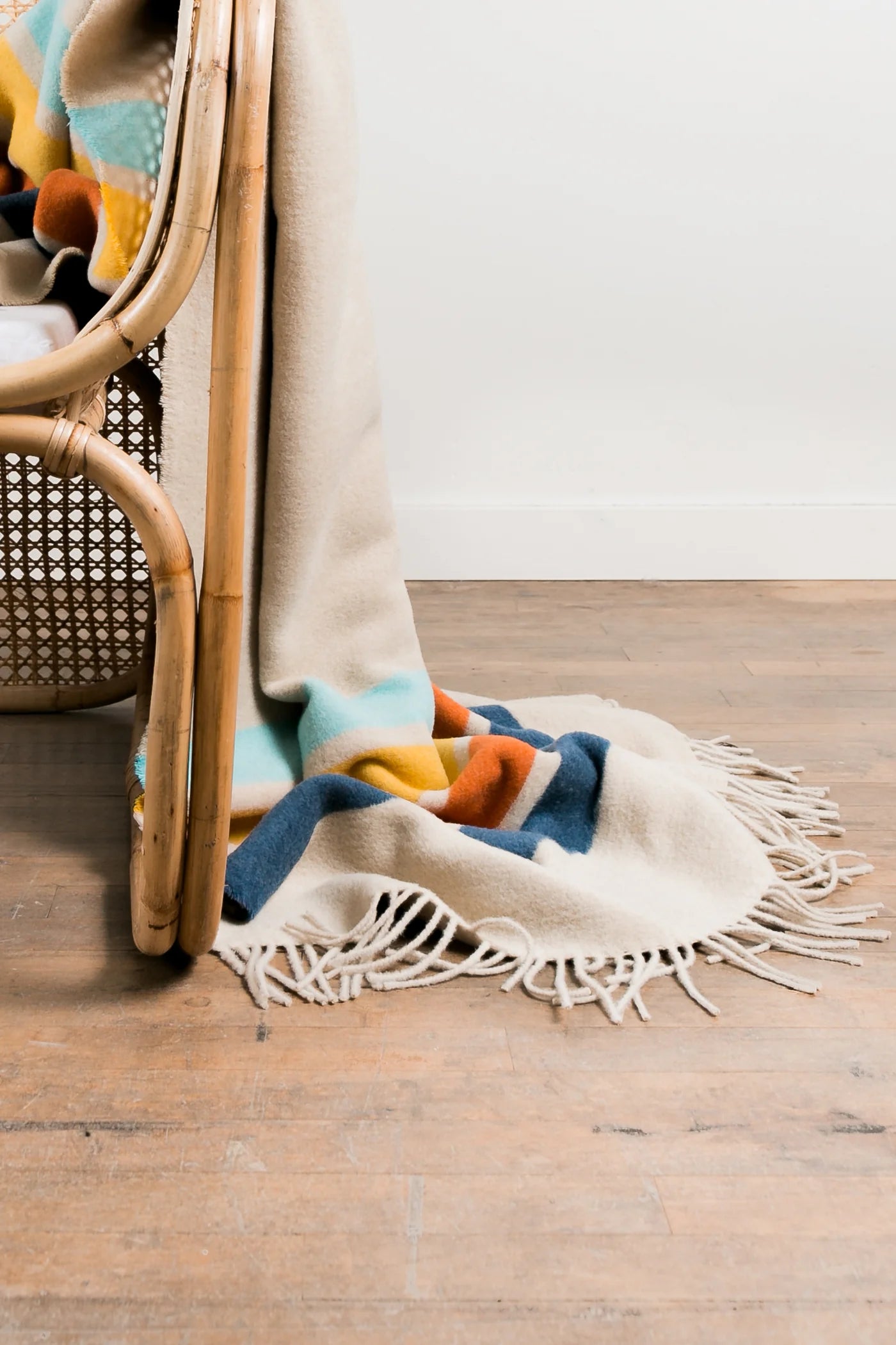 The Sackcloth & Ashes/Camp Coast blanket by Sackcloth & Ashes, cream-colored with colorful geometric patterns and fringe, drapes over a wicker chair onto a wooden floor against a plain white wall.