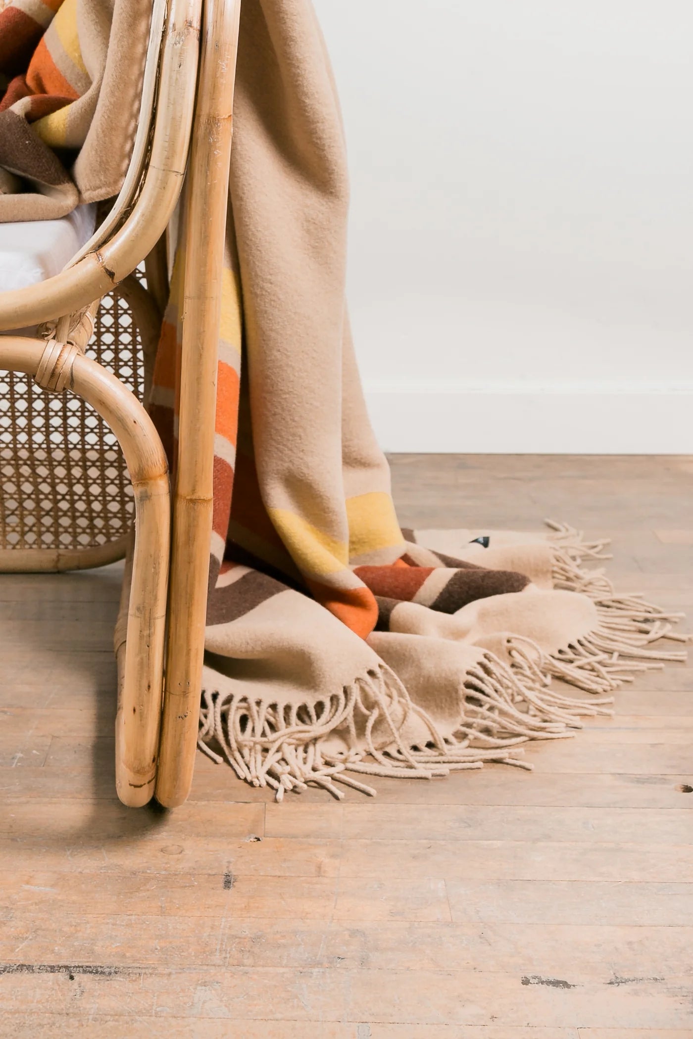 The Sackcloth & Ashes/Desert Camp blanket by Sackcloth & Ashes, with its colorful fringe, drapes over a woven wooden chair on a light wood floor—an inviting accent for any room.