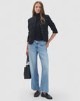 A woman stands against a plain background wearing a black buttoned jacket, black turtleneck, Rag & Bone ftwt saige ankle straight jeans in light blue, and black loafers, holding a black handbag at her side.