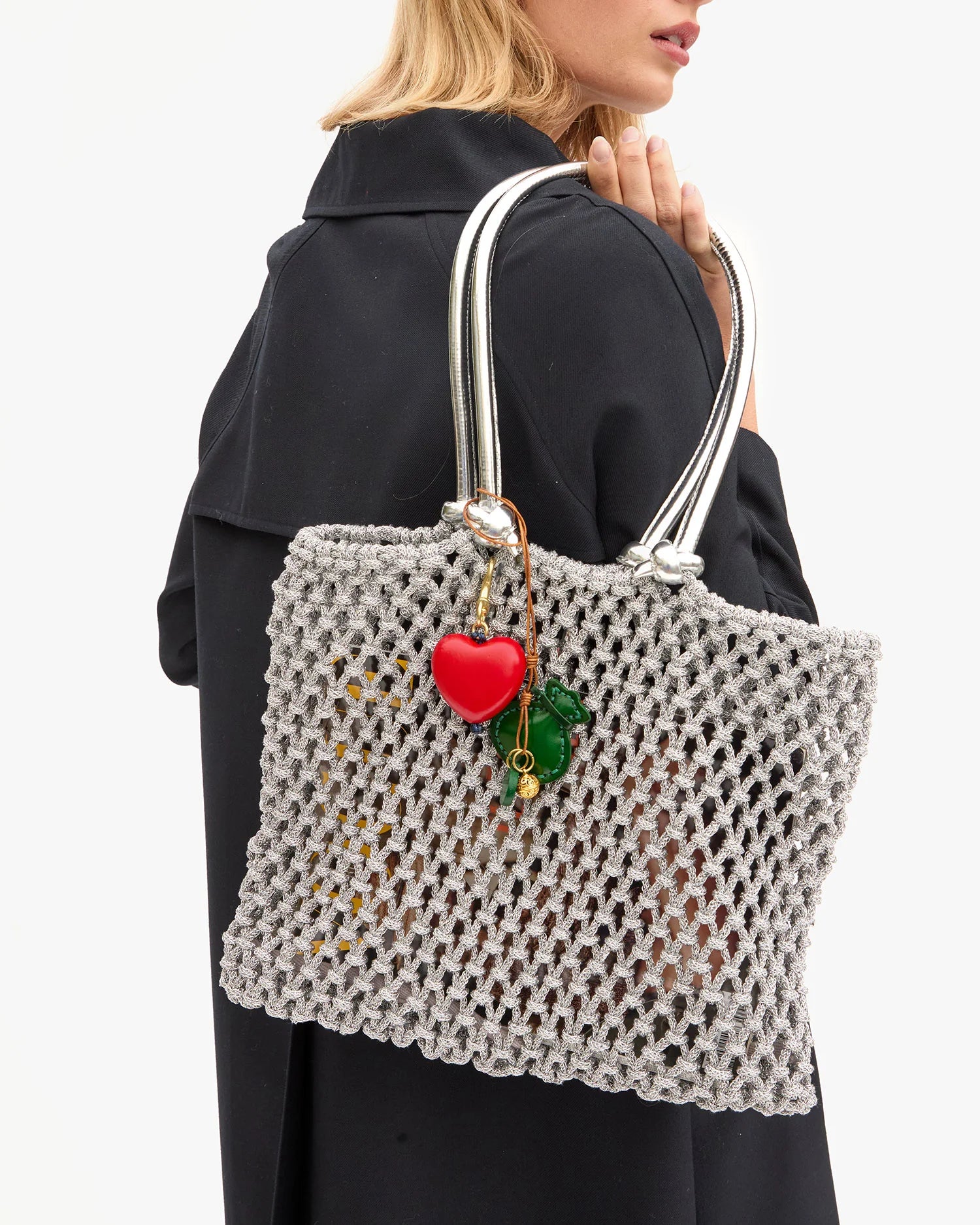 A woman with blonde hair in a black coat carries a silver mesh handbag over her shoulder, adorned with the Clare Vivier Grande Heart Fob /Poppy keychain and green pepper charms.