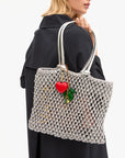 A woman with blonde hair in a black coat carries a silver mesh handbag over her shoulder, adorned with the Clare Vivier Grande Heart Fob /Poppy keychain and green pepper charms.