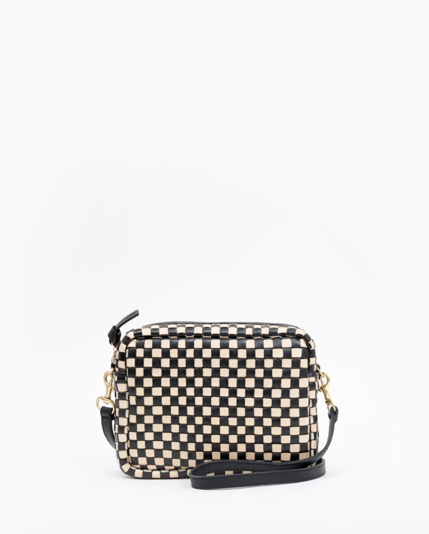 A small rectangular crossbody bag with a black and white checkered pattern. The handwoven leather Midi Sack by Clare Vivier features a black adjustable strap attached to gold hardware and a zippered top. The background is plain white.