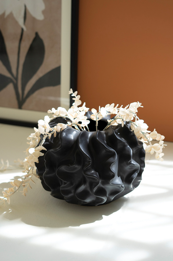A Black Coral Ceramic Vase adorned with white flowers sits on a sunny ledge in an Arizona-style bungalow, against a backdrop of a painting with floral motifs and an orange wall.
