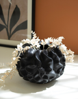 A Black Coral Ceramic Vase adorned with white flowers sits on a sunny ledge in an Arizona-style bungalow, against a backdrop of a painting with floral motifs and an orange wall.