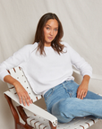 A woman sits relaxed in a white chair in her bungalow, wearing a Perfectwhitetee Ziggy Reverse Fleece LS Shrunken Crewneck Sweatshirt and blue jeans. She gazes at the camera with a composed expression, her long brown hair loosely framing her face.