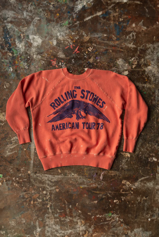 A vintage "The Rolling Stones American Tour '78" Shrunken Sweat in rust orange color, displayed on a textured, multicolored, worn-out background in a Scottsdale Arizona bungalow by Made Worn.