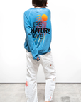 A person with short hair stands facing away, showcasing unisex fashion with the LNLSUN SprYum Biggie Raglan from Free City (sparrow, LLC), a blue oversized sweatshirt featuring a sun graphic and the text "If Nature Loves." Paired with white pants adorned with red patches and black slippers, they stand against a plain white wall.