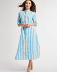 A woman with long hair wearing a blue and white striped Sadie Long Caftan by Mille stands barefoot in a Scottsdale, Arizona studio, her hair tossed mid-motion, looking directly at the camera.