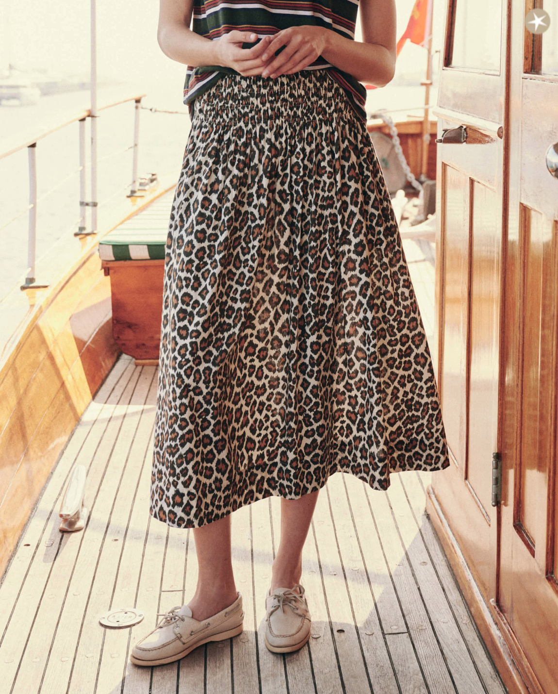 Person standing on a wooden deck of a boat, wearing The Viola Skirt by The Great Inc. with a smocked waistband and white boat shoes. They are holding an object in both hands at waist height. Sunlight filters through, casting shadows on the deck.