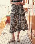 Person standing on a wooden deck of a boat, wearing The Viola Skirt by The Great Inc. with a smocked waistband and white boat shoes. They are holding an object in both hands at waist height. Sunlight filters through, casting shadows on the deck.