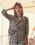 A woman stands outdoors, wearing The Post Top by The Great Inc. with tailored shirt cuffs. She has her left hand resting on her head, looking directly at the camera. The background suggests a sunny day on a porch or deck.