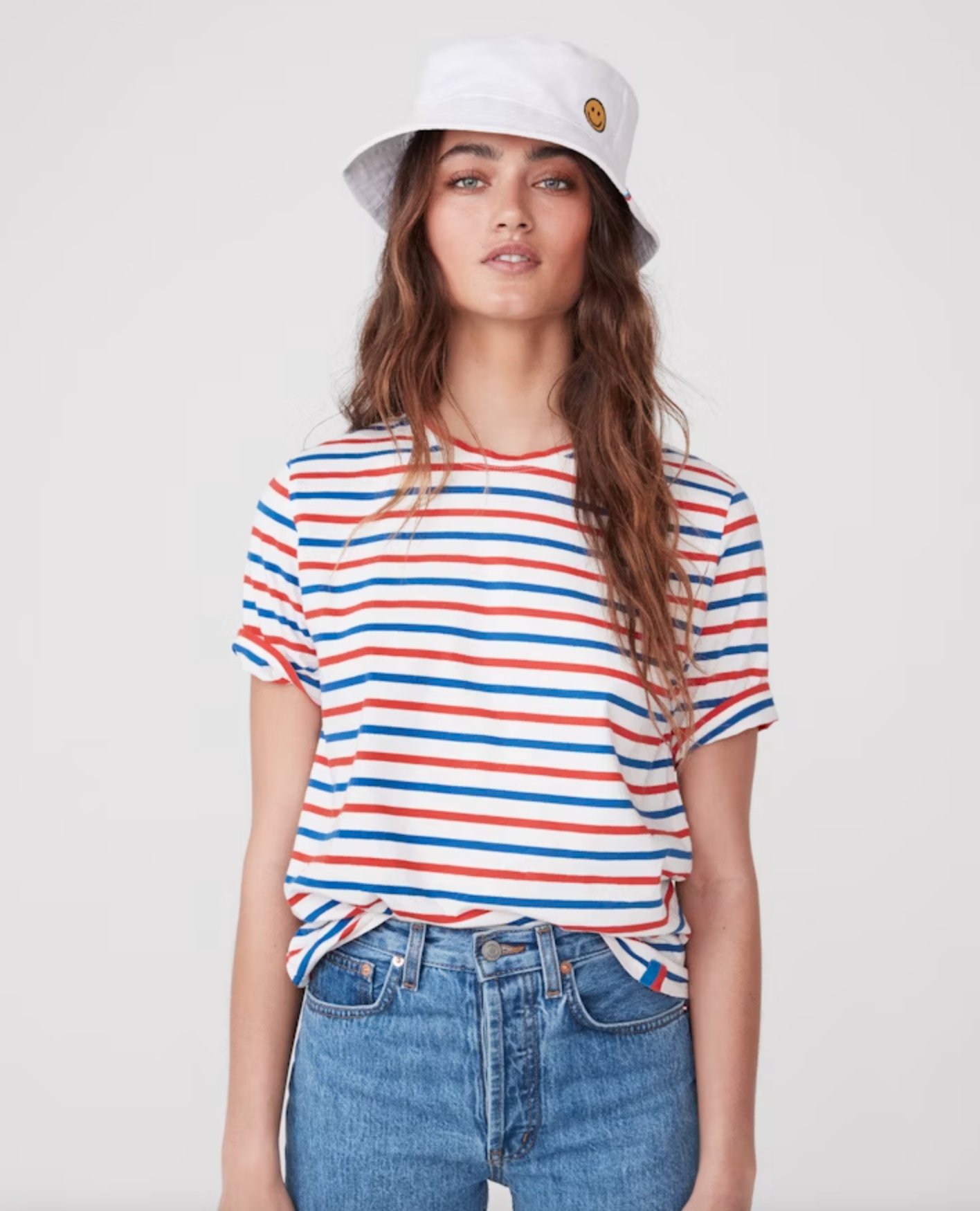 A woman with long brown hair is wearing a white bucket hat with a small smiley face patch, The Modern from Kule featuring red, blue, and white horizontal stripes, and blue jeans. She is standing against a plain white background, looking directly at the camera.