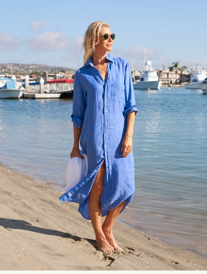 A person wearing a classic Rory Shirtdress by Frank & Eileen and sunglasses walks barefoot along a sandy beach, holding a white hat. In the background, there is a marina with boats docked and buildings along the waterfront.