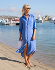 A person in a light blue, 100% linen Frank & Eileen Rory Shirtdress S24 stands barefoot on a sandy beach, wearing sunglasses and holding a white hat. The loose-fit, maxi-length dress flows gracefully as boats are visible in the water and a coastal town is seen in the background under a clear blue sky.