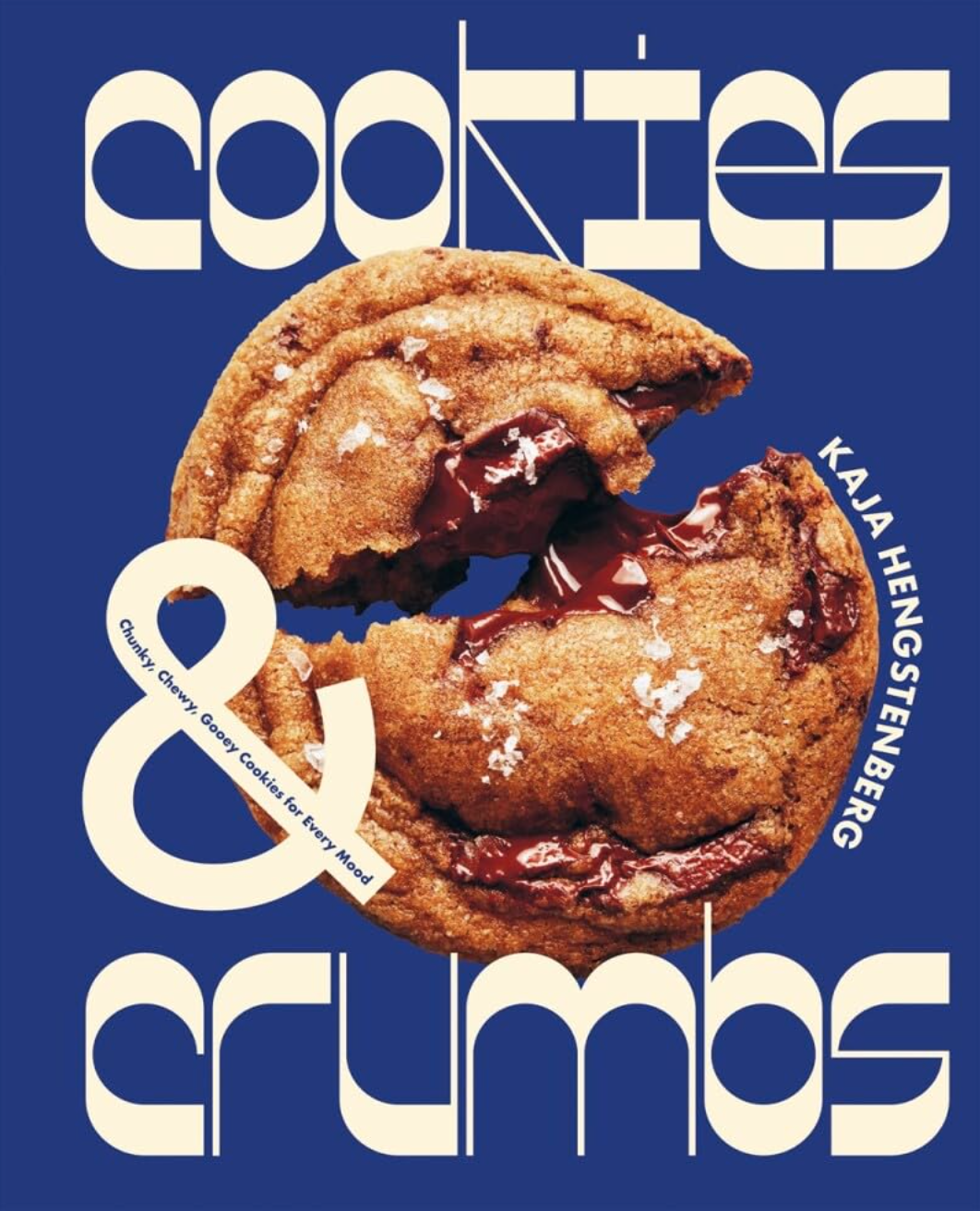 A book cover titled "Cookies & Crumbs" by Kaja Hengstenberg, featuring a close-up of a chocolate chip cookie with melted chocolate on a blue background. The title is displayed in a bold, modern font. The subheading reads "Classic, Crunchy, Gooey Cookies and Baking Recipes for Every Mood." This product from Hachette Book Group is perfect for baking enthusiasts looking to explore new recipes.