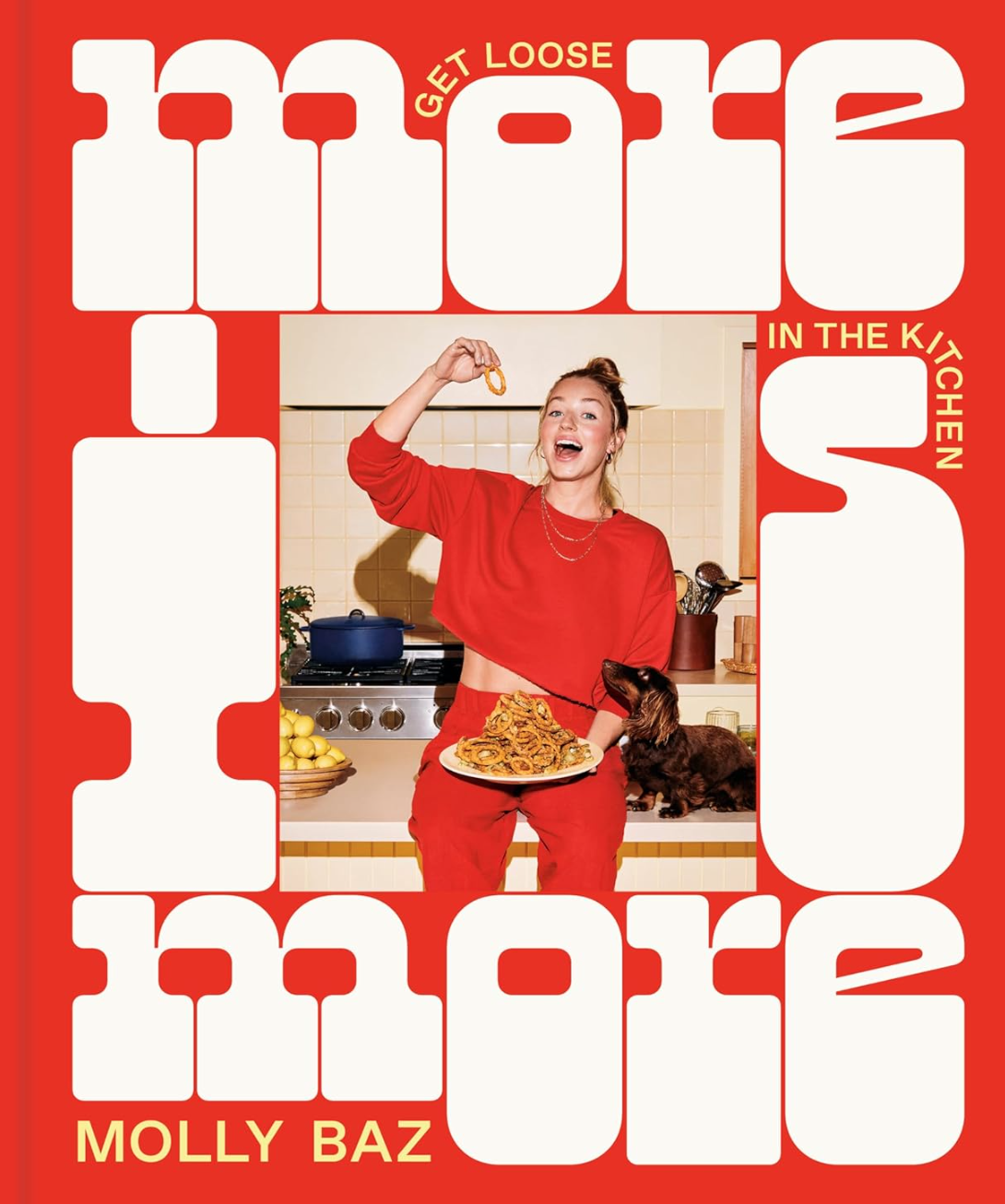 A cookbook cover titled "More Is More: Get Loose in the Kitchen." It features a woman in a red outfit holding a plate of food and interacting with a dog. The background shows a kitchen with various ingredients and utensils. The Random House cookbook, More is More, hailed as a NEW YORK TIMES BESTSELLER, is full of bold flavorful recipes.