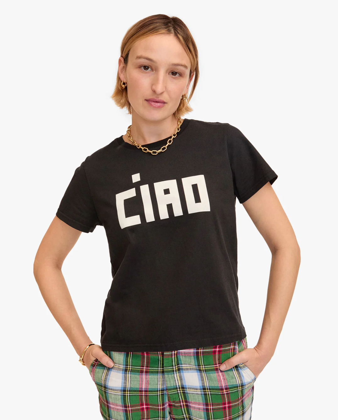 A person with short, light brown hair is wearing a black Clare Vivier Classic Tee made from 100% cotton, featuring the word "Ciao" in bold, white letters. They have gold hoop earrings, a gold chain necklace, and gold bracelets. Additionally, they are wearing plaid pants with green, red, and white stripes.